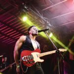 Nigerian artist Jarell Ebuka performing live on stage while singing into a microphone and playing a red electric guitar under vibrant yellow and red stage lights.