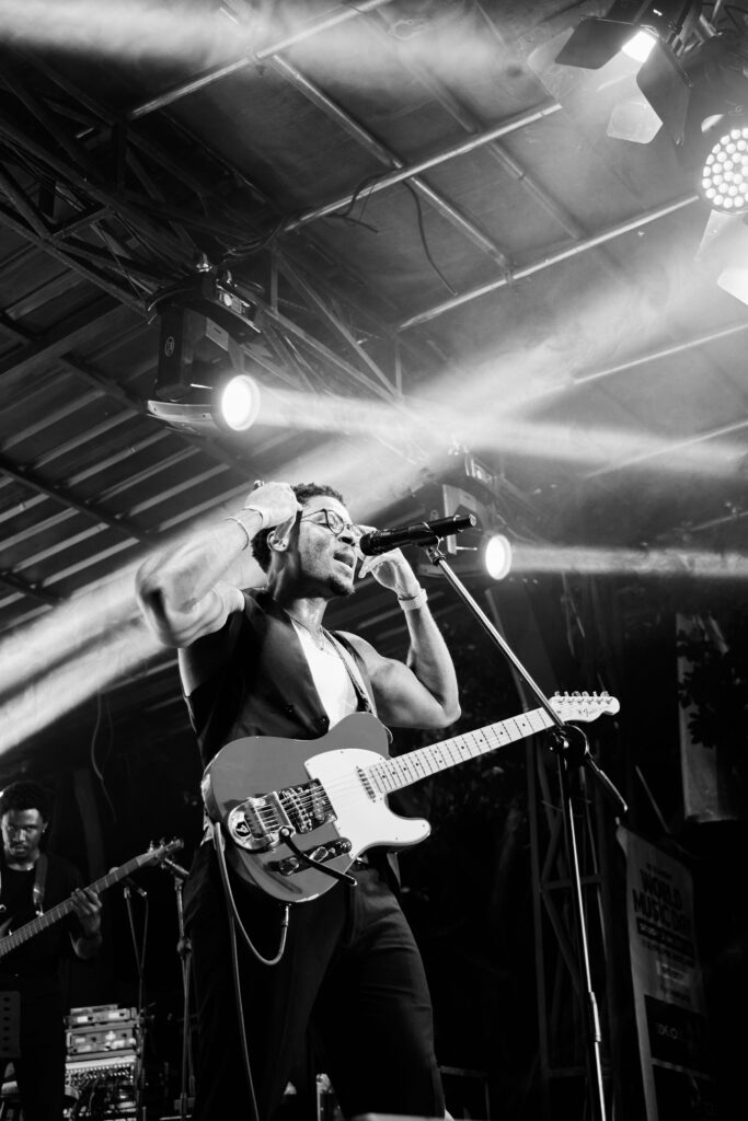 this is a Black and white concert photography of Nigerian artist Jarell Ebuka performing live, singing into a microphone and playing an electric guitar under dramatic stage lights.