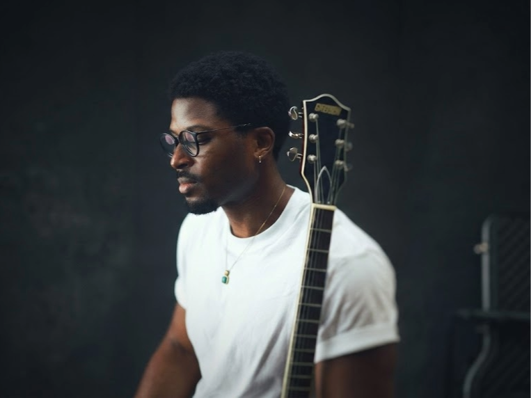 Portrait of Nigerian artist Jarell Ebuka wearing a white t-shirt and glasses, holding a Gretsch electric guitar, posing against a dark background.