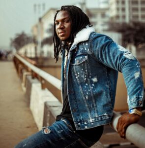 Nigerian folk musician Francis Atela in a distressed denim jacket with a fleece collar, leaning on a bridge railing during a promotional outdoor photoshoot.