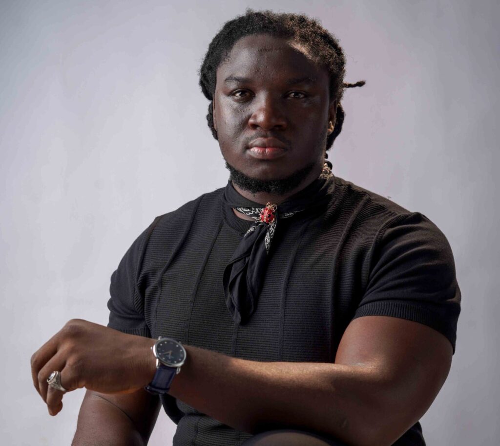 Promotional portrait of Nigerian folk artist Francis Atela wearing a black short-sleeved shirt and neck bandana, looking thoughtfully into the camera.