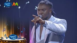 Francis Atela performing live on stage during Nigerian Idol Season 6, holding a microphone and wearing a light blue suit