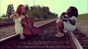 Blessing Tangban playing an acoustic guitar alongside a cellist on outdoor train tracks in the official Nowhere Girl music video.