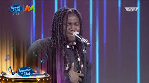 Francis Atela singing passionately on Nigerian Idol Season 6 with his hand on his chest, wearing a dark outfit and traditional beads.
