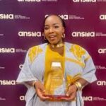 Blessing Tangban smiling and holding her clear glass trophy in front of a purple ANSMA Awards step-and-repeat backdrop.